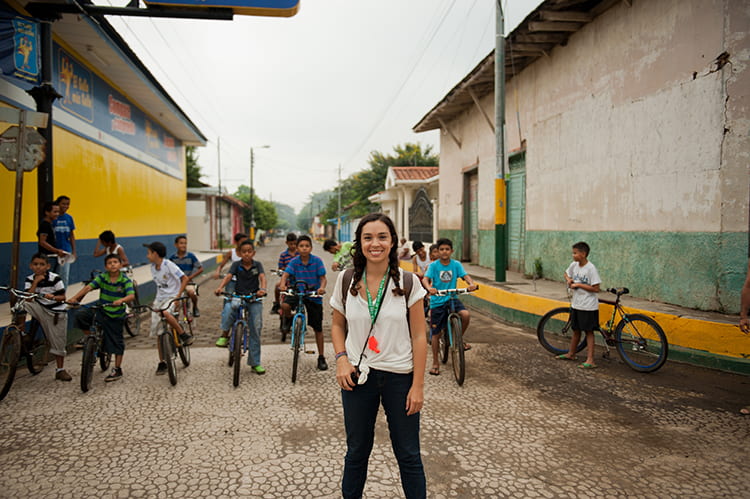A young woman stands in a street in Nicaragua, a group of boys on bicycles are in a line behind her. The woman is smiling and wearing a white shirt and dark jeans. She has a camera hanging around her neck. The boys are all wearing different colored shirts and pants. They are all smiling and at the camera. The street is made of cobblestones and there are buildings on either side of the street. The buildings are made of concrete and have brightly colored walls. 