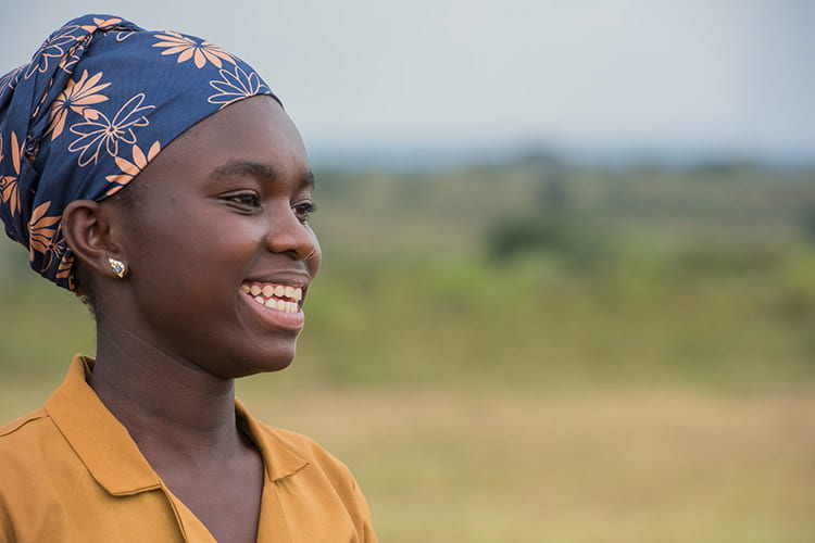 Smiling young Cameroonian female. She has a gold earring and a blue head scarf with orange hued flowers printed into the fabric. She is wearing an orange shirt.